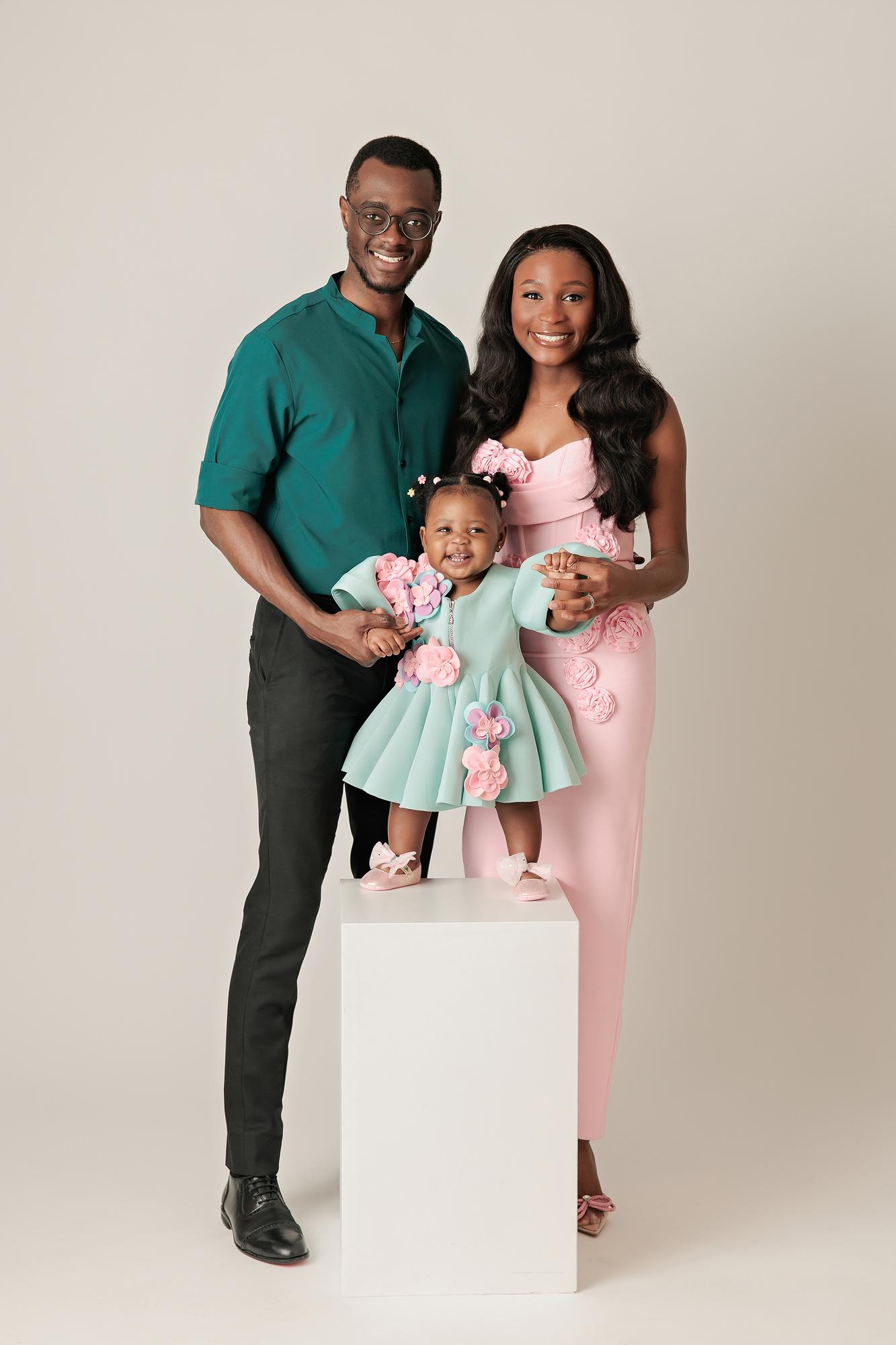 Family photography Calgary studio: Joyful family of three — father in a teal shirt and mother in a blush pink 3D floral gown — with their smiling baby girl in a mint and pink floral dress standing on a white plinth, light grey background